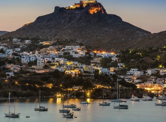 Παραθαλάσσιος οικισμός στη Λέρο με λευκά σπίτια και ιστιοπλοϊκά αγκυροβολημένα στο λιμάνι, με φωτισμένο κάστρο στην κορυφή λόφου κατά το σούρουπο.