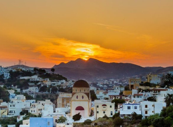Παραδοσιακός οικισμός Λέρου με λευκά και παστέλ κτίρια, εκκλησία με κόκκινο τρούλο στο κέντρο και ηλιοβασίλεμα πίσω από βουνά.